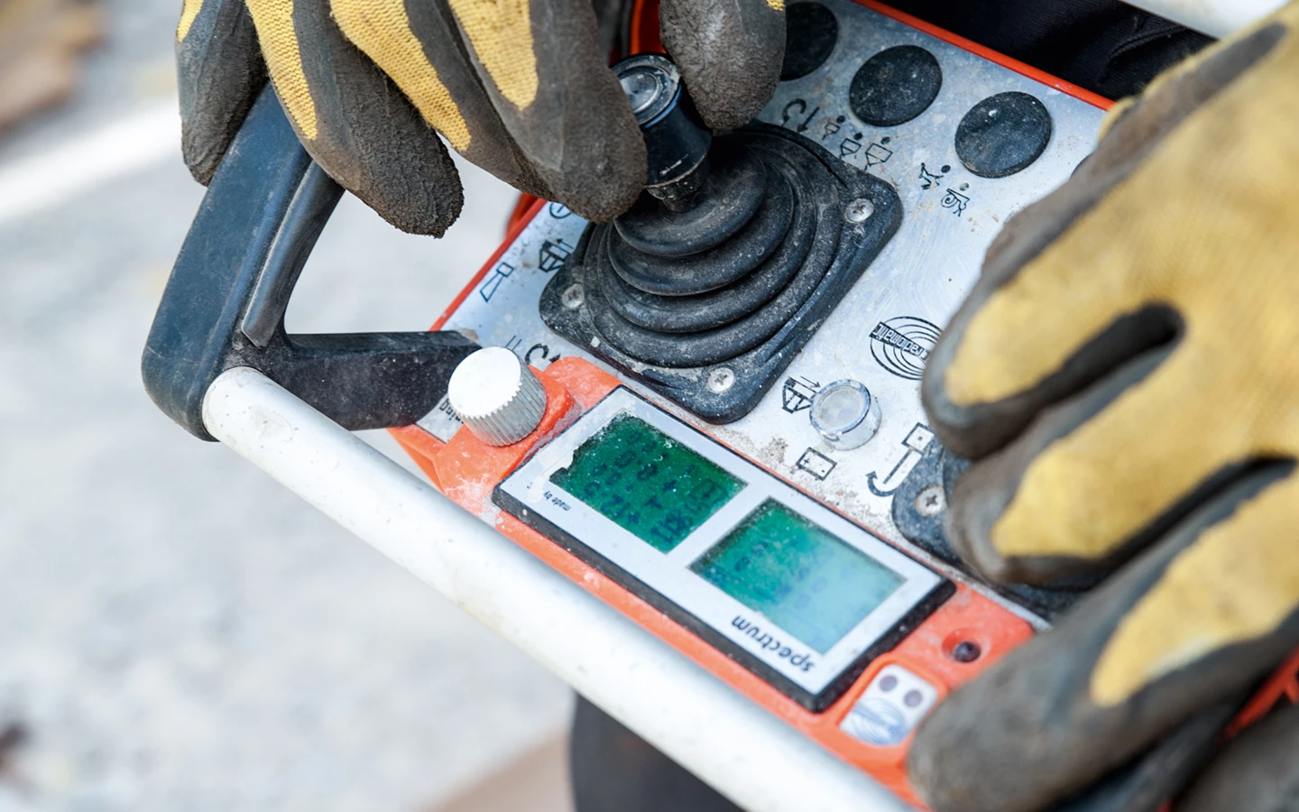 Arbeiter in gelben Schutzhandschuhen bedient ein orange-graues Funkfernsteuerungsgerät mit zwei Bildschirmen und einem Joystick (Baustelle, Hochbau)