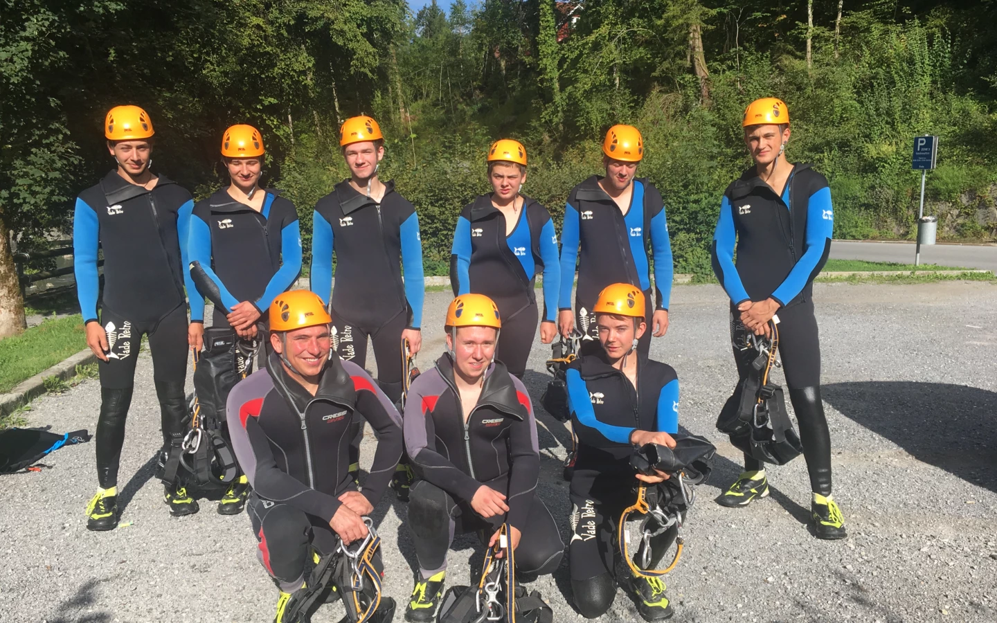 Eine Gruppe von neun jungen Erwachsenen in Neoprenanzügen und orangefarbenen Helmen, bereit für Outdoor-Aktivitäten (Lehrlingen, Rafting, Ausrüstung).