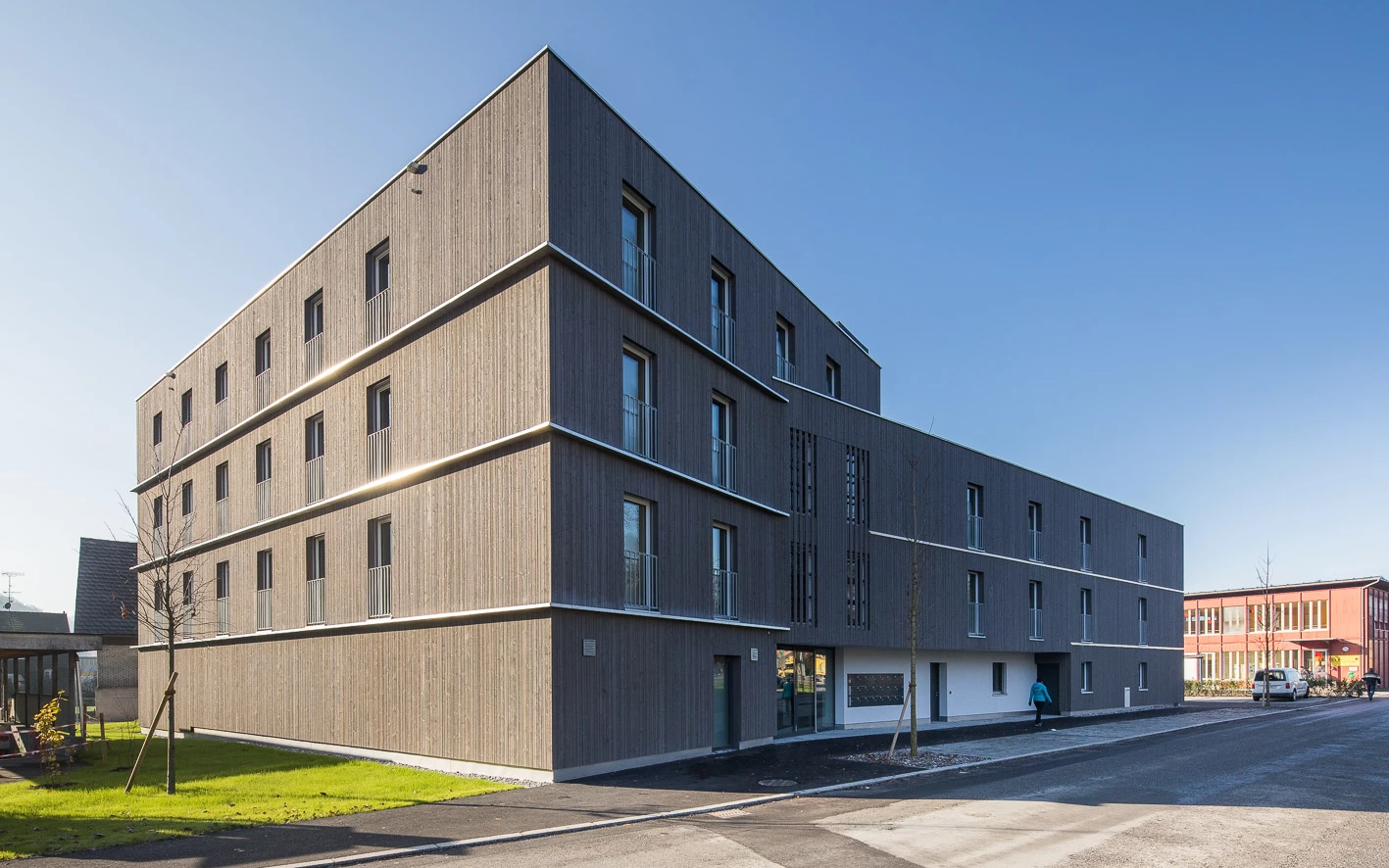 Modernes, mehrstöckiges Wohngebäude mit Holzfassade und Balkonen unter blauem Himmel, entlang einer Straße mit Grünflächen (Froschweg).
