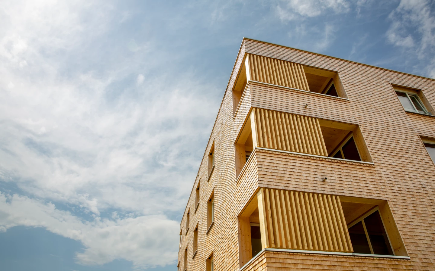 Holzfassade eines modernen Gebäudes mit vertikalen Holzlamellen und hellen Fenstern unter bewölktem Himmel (Hellbrunnenstraße).
