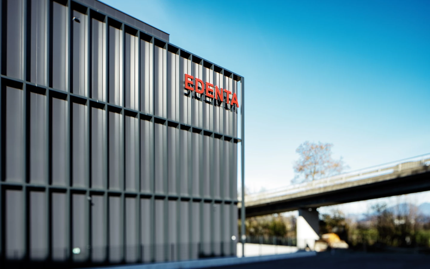 Modernes Gebäude mit dem roten Logo "EDENTA" an der Fassade unter blauem Himmel, einer Brücke und Bäumen im Hintergrund.