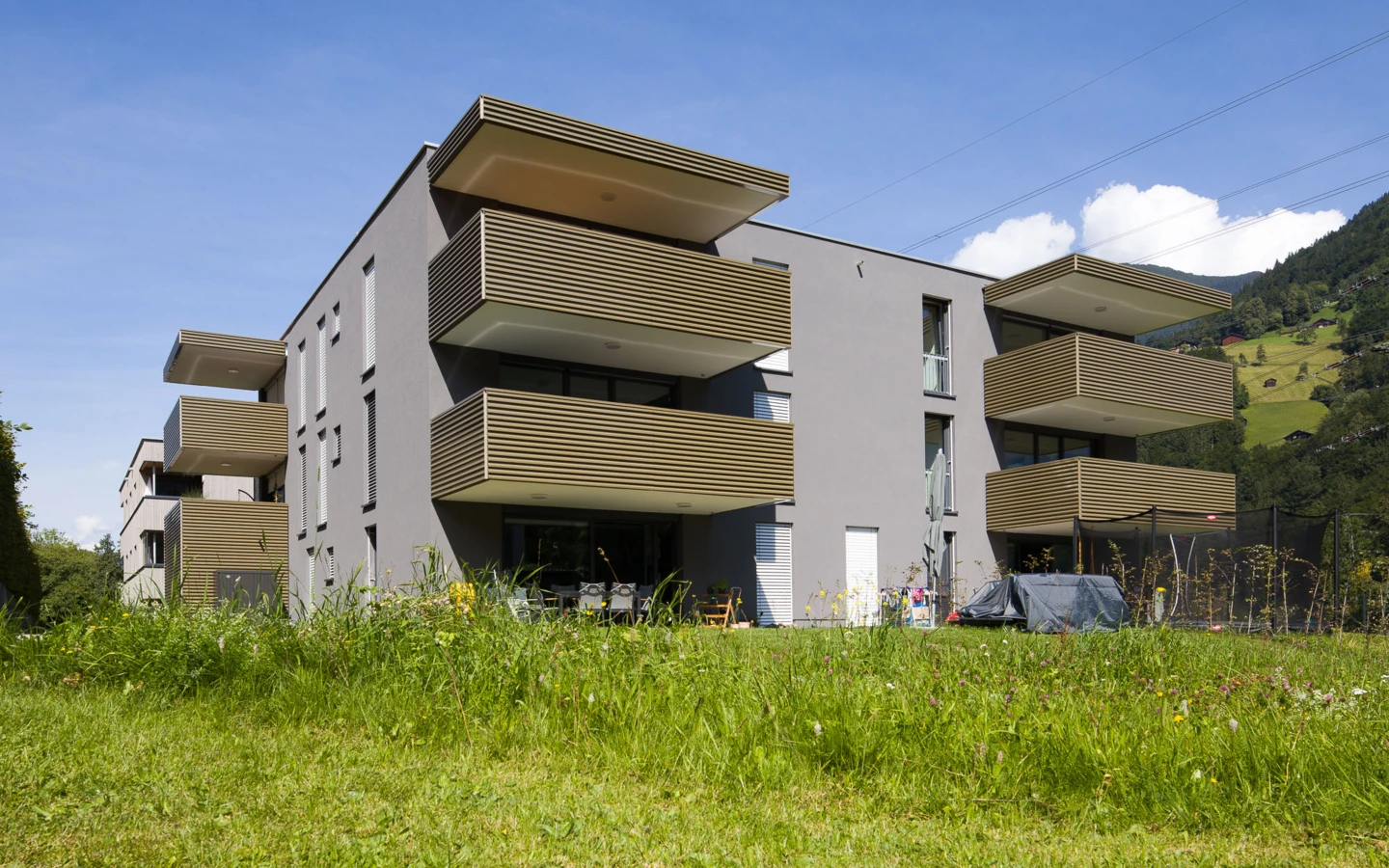 Modernes Wohnhaus in Vandans mit grauer Fassade, hellen Holzlamellenbalkonen auf einer grünen Wiese unter blauem Himmel (Bahnhofstraße, Wohnanlage, Bahnhofsstrasse).