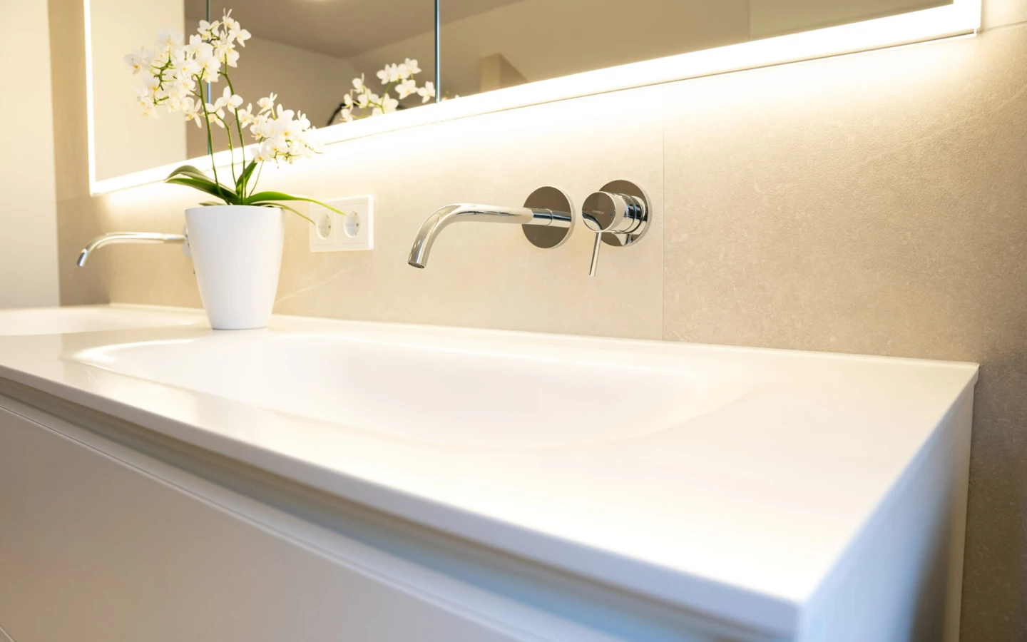 Modernes Badezimmer mit weißem Waschtisch, Wandarmatur, beleuchtetem Spiegel und einer weißen Orchidee in einem Topf (Generalsanierung, Bad).