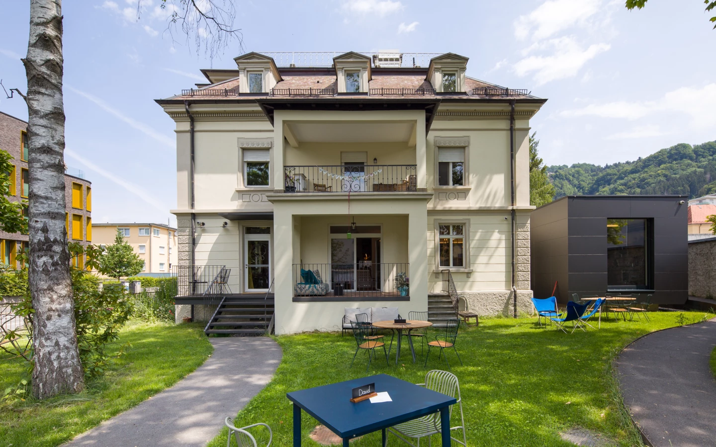 Historisches helles Mehrfamilienhaus mit Vorgarten und Terrassen, ausgestattet mit Tischen und Stühlen auf grünem Rasen (Wunderkind, Backstube).