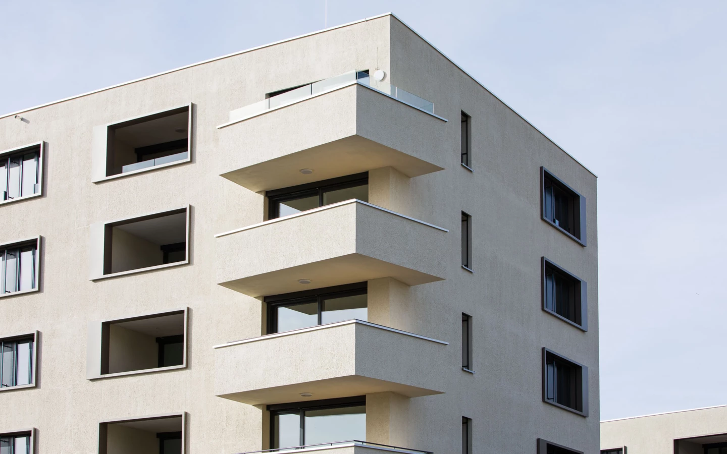 Moderne, hellbeige Fassade eines mehrstöckigen Wohngebäudes mit dunklen Fenstern und gestuften Balkonen an der Ecke (Bäumleareal).