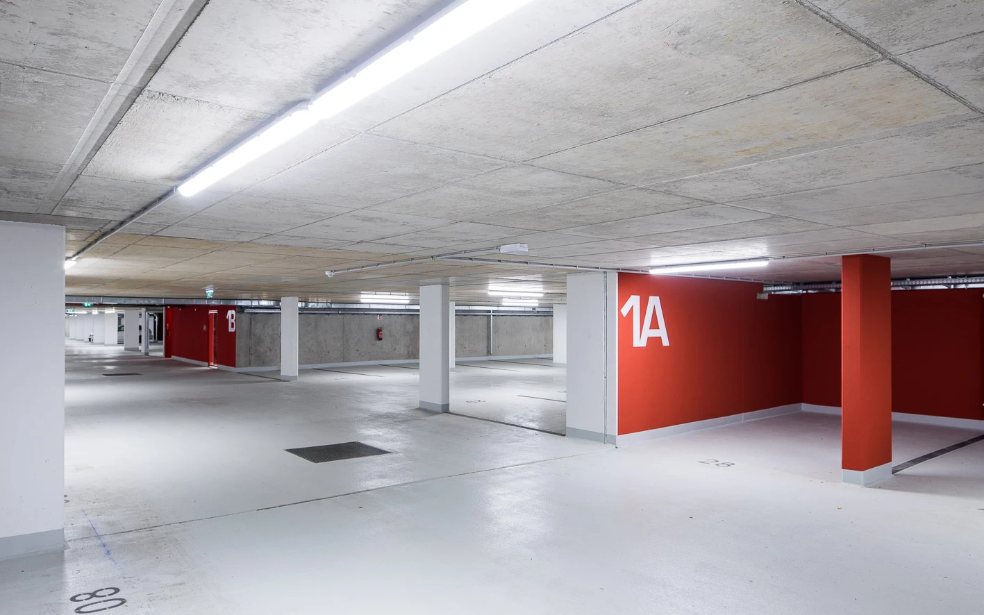 Leere Tiefgarage mit Betondecke, weiß gestrichenen Säulen und leuchtend roten Wänden, die mit den Zahlen 1A und 1B gekennzeichnet sind (Baeumlequartier).