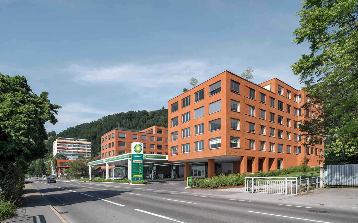 Zwei moderne orangefarbene Backsteingebäude neben einer BP-Tankstelle an einer Straße in Lochau, mit bewaldeten Hügeln im Hintergrund (Seeblick).