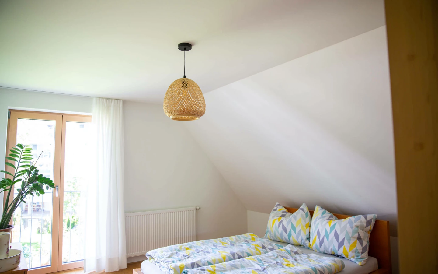 Ein helles Schlafzimmer in einem Einfamilienhaus in Dornbirn mit Holzbett, bunter Bettwäsche und Bambuslampe (Generalsanierung).