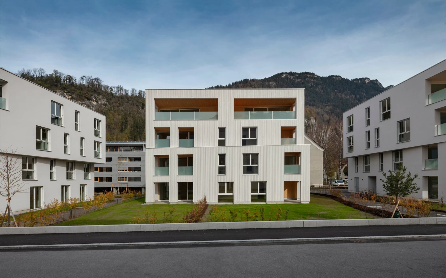 Moderne Wohnanlage Zürcherstrasse Bludenz mit weiß verputzten Fassaden, Holzverkleidungen und Balkonen vor Grünflächen und Bergen (Holzbau).