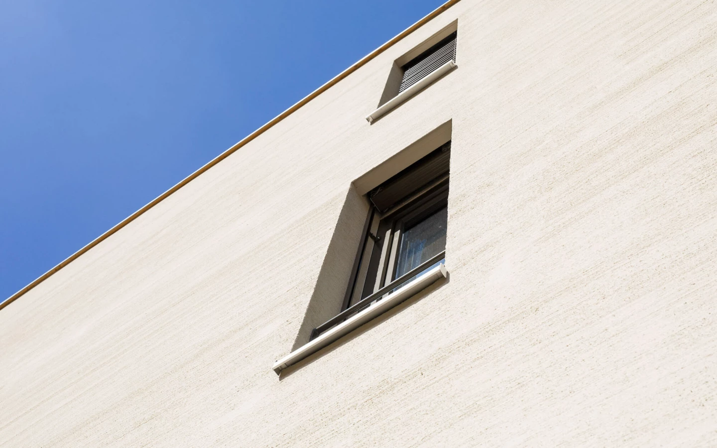 Ansicht einer hellbeigen Fassade einer Wohnanlage mit zwei Fenstern und Rollos vor klarem blauem Himmel (Feldgasse, Dornbirn).