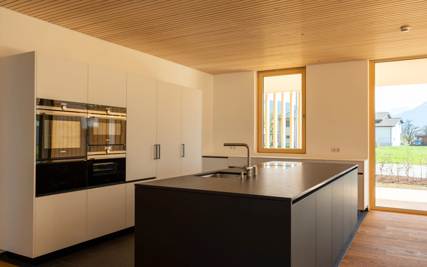 Moderne Küche mit integrierten Siemens-Geräten, dunkler Kücheninsel mit Spülbecken und hellbrauner Holzdecke. Fenster bieten Blick ins Grüne (Wolfurt, Dieter).