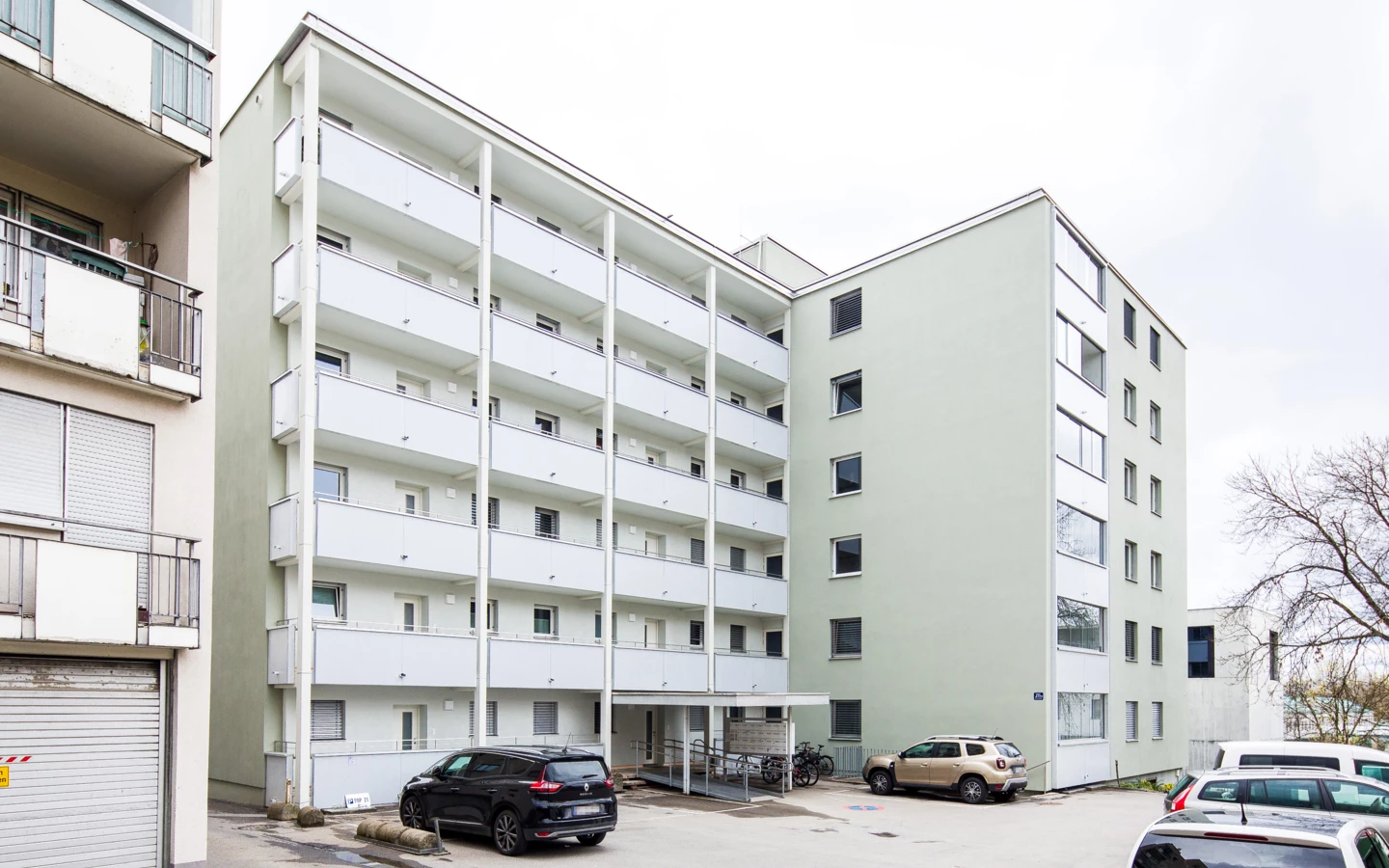 Mehrgeschossiges Wohngebäude mit hellgrüner und weißer Fassade sowie Balkonen und einem Parkplatz davor. Mehrere Autos sind geparkt (Römerstraße).