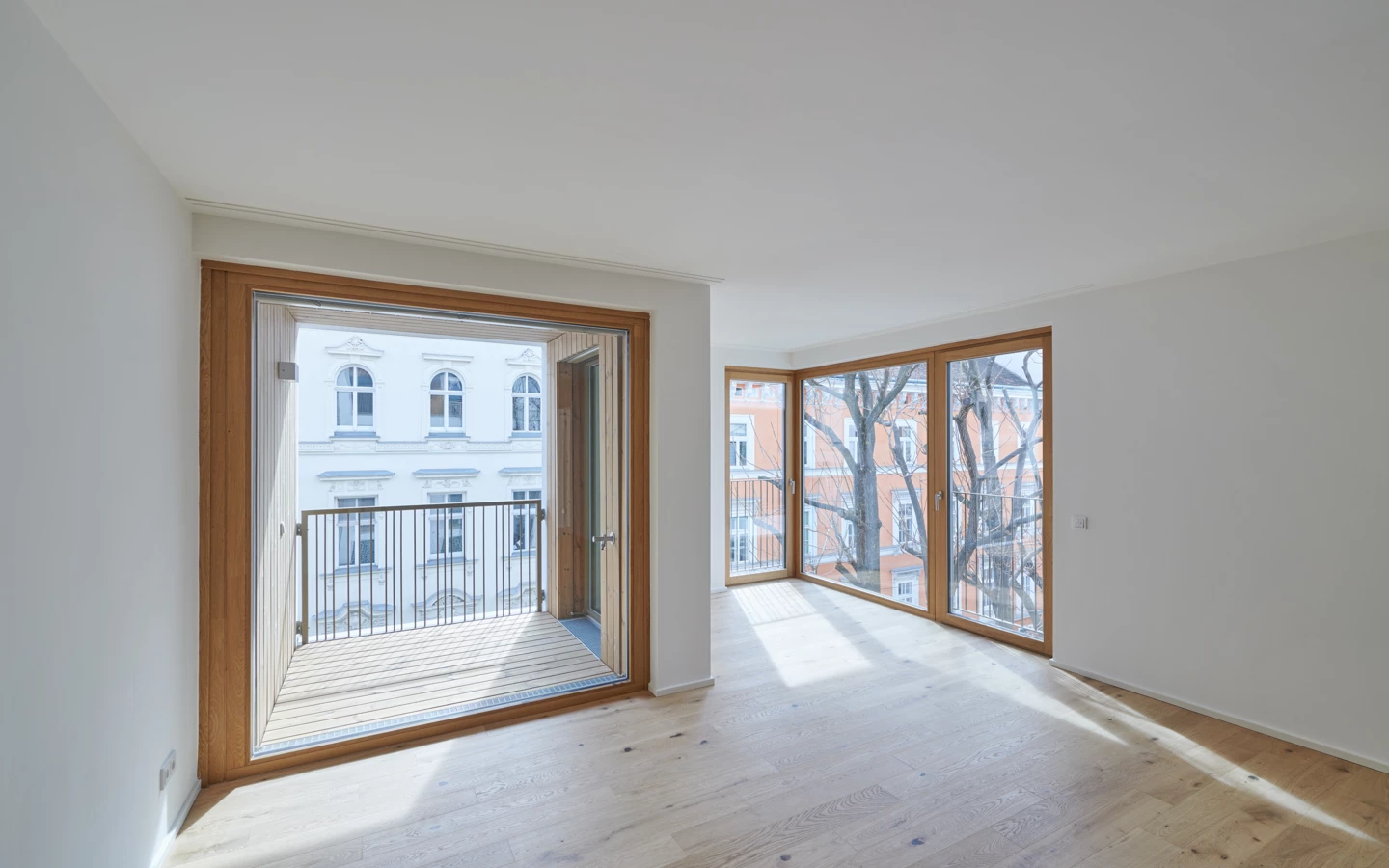 Heller Raum mit Parkettboden und großen Fenstern sowie einer Balkontür, die auf einen Balkon mit Blick auf ein weißes Gebäude führt (Apollogasse).