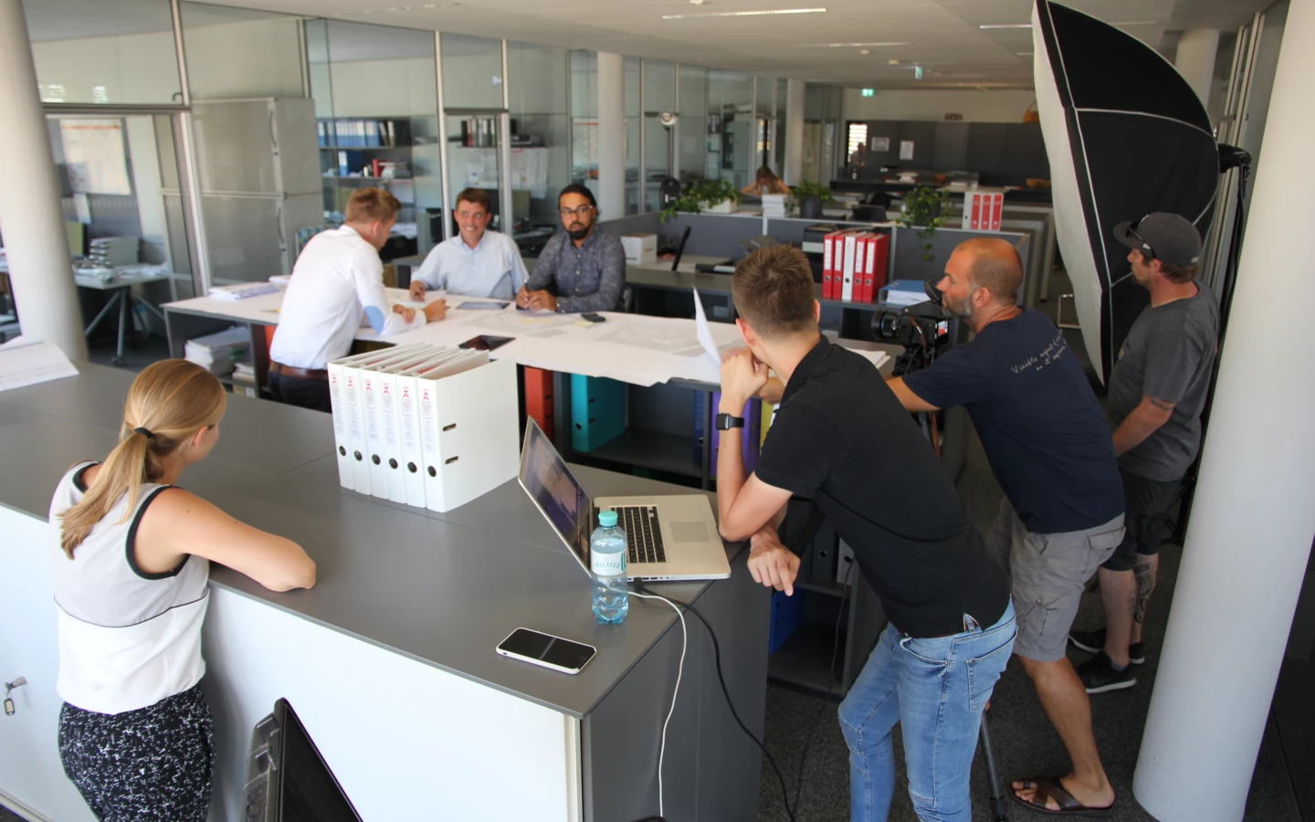 Geschäftsleute sitzen an einem Tisch, während ein Kamerateam in einem modernen Rhomberg Bau Büro filmt.