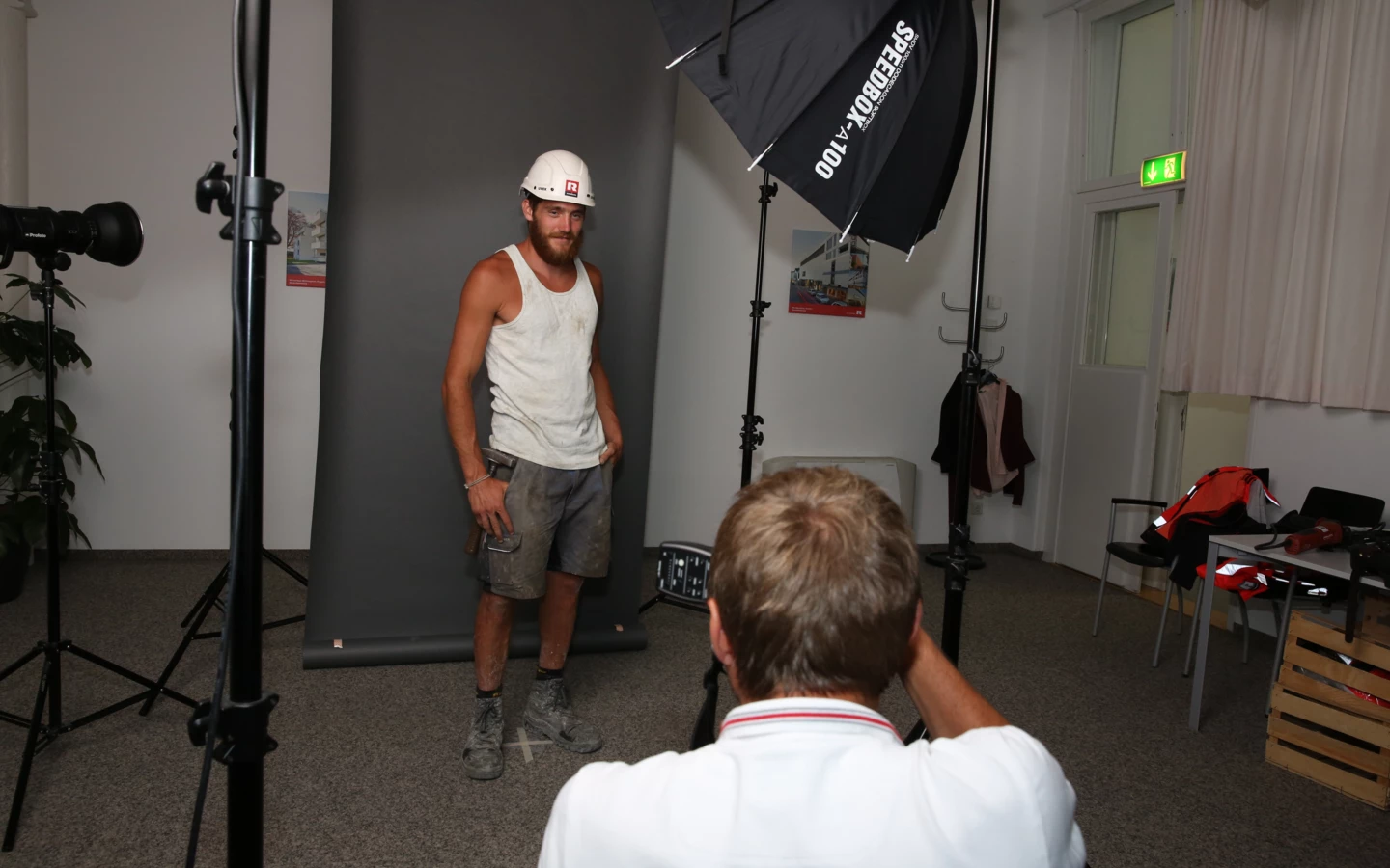 Ein bärtiger Bauarbeiter mit Helm posiert vor einer grauen Leinwand für ein Fotoshooting, während ein Fotograf im Vordergrund steht.