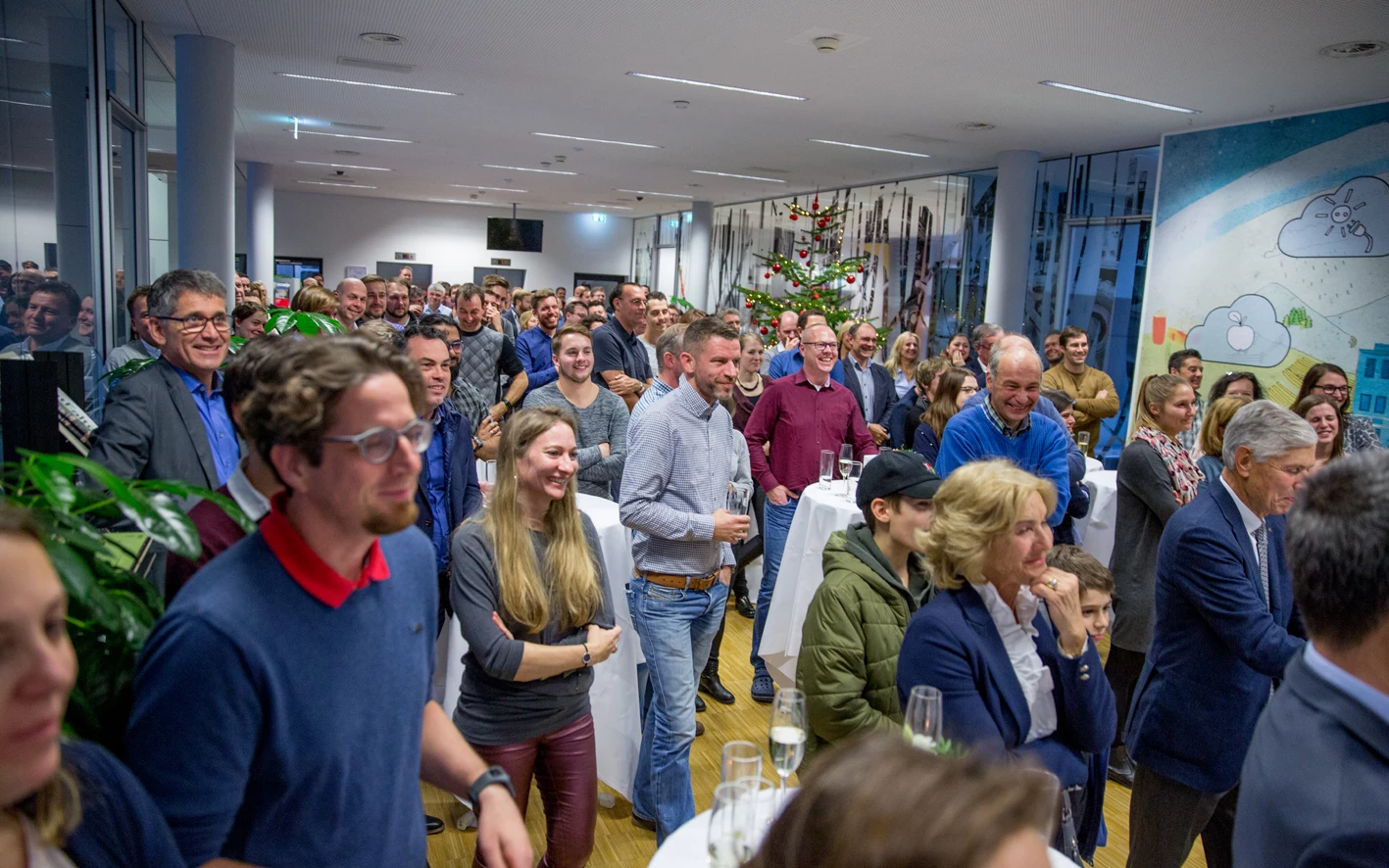 Eine große Gruppe lachender Menschen, die eine Präsentation oder Veranstaltung genießen, mit einem Weihnachtsbaum im Hintergrund (Hubert, Geburtstag).