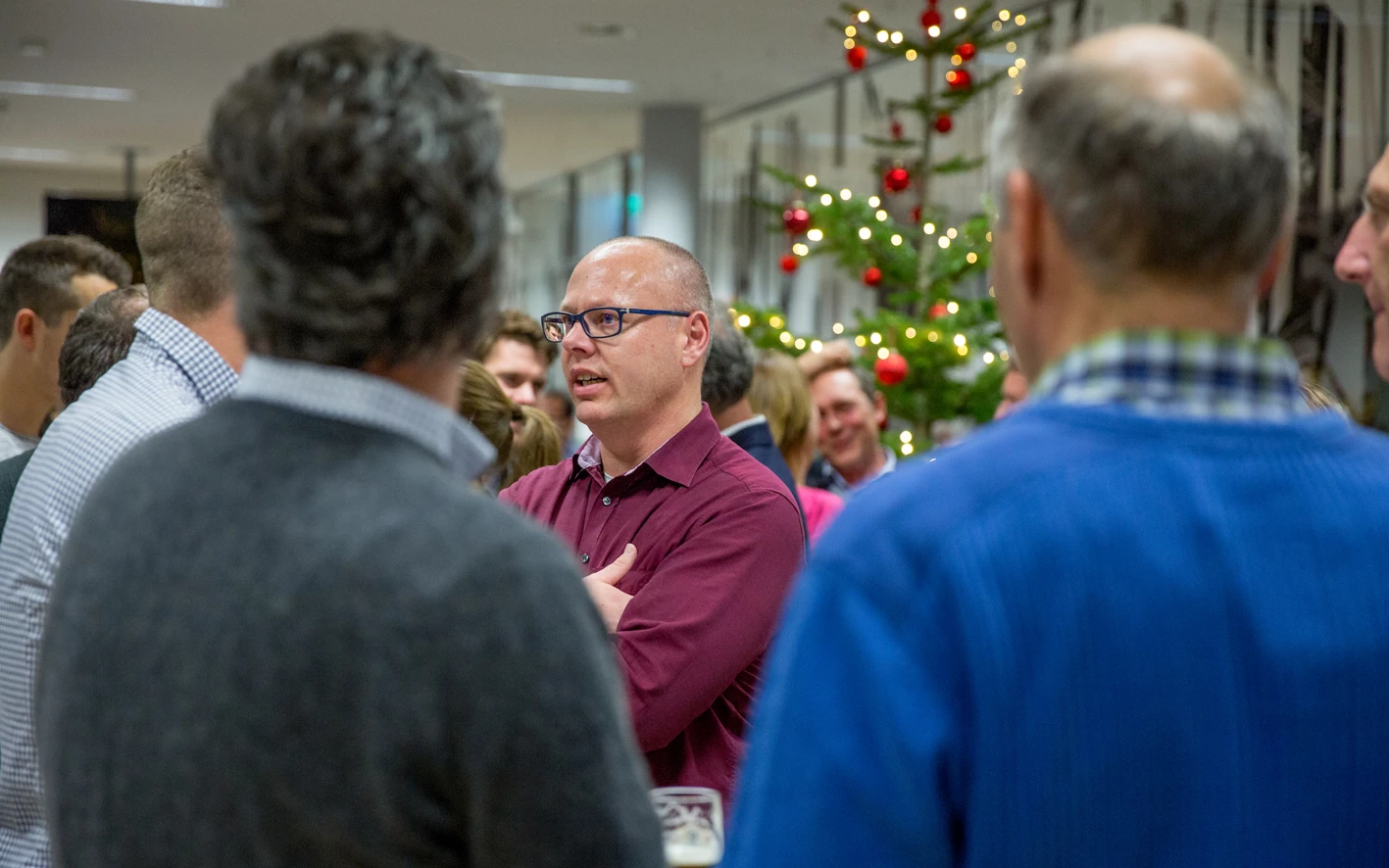 Ein Mann mit Brille und weinrotem Hemd spricht in einer Gruppe von Menschen, im Hintergrund ein Weihnachtsbaum (Hubert, Geburtstag).
