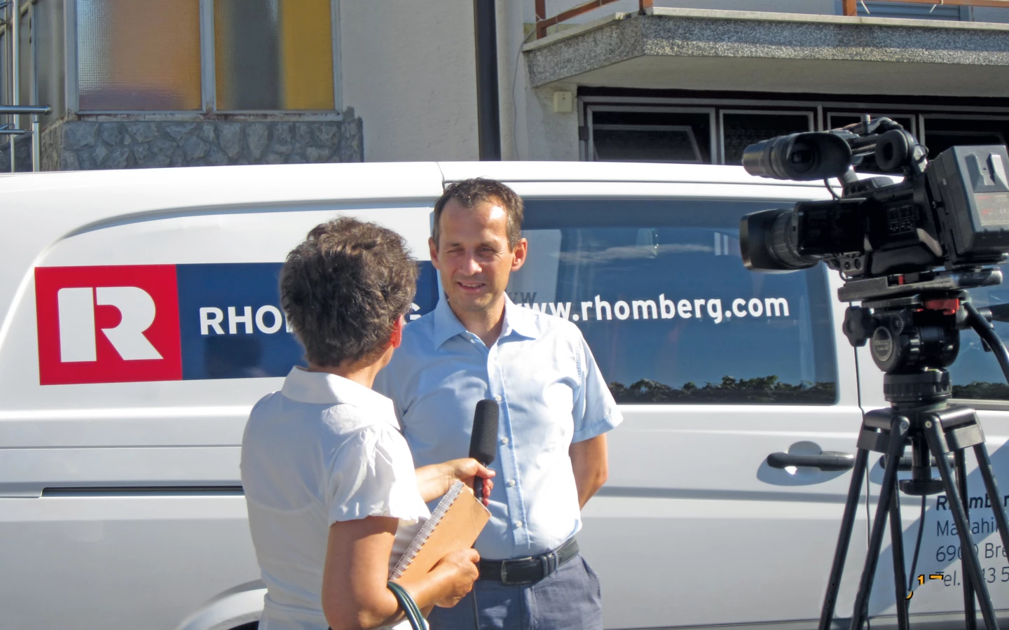A man in a light blue shirt is interviewed by a woman holding a microphone and notebook, beside a Rhomberg Bau van and a camera on a tripod (pensioners)