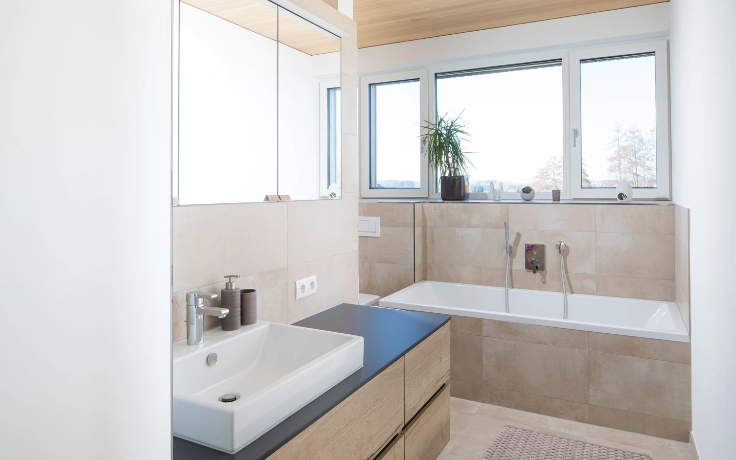 Modernes Badezimmer mit weißer Badewanne, Eckwaschbecken auf Holzunterschrank und einem großen Fenster (Hohenweiler)