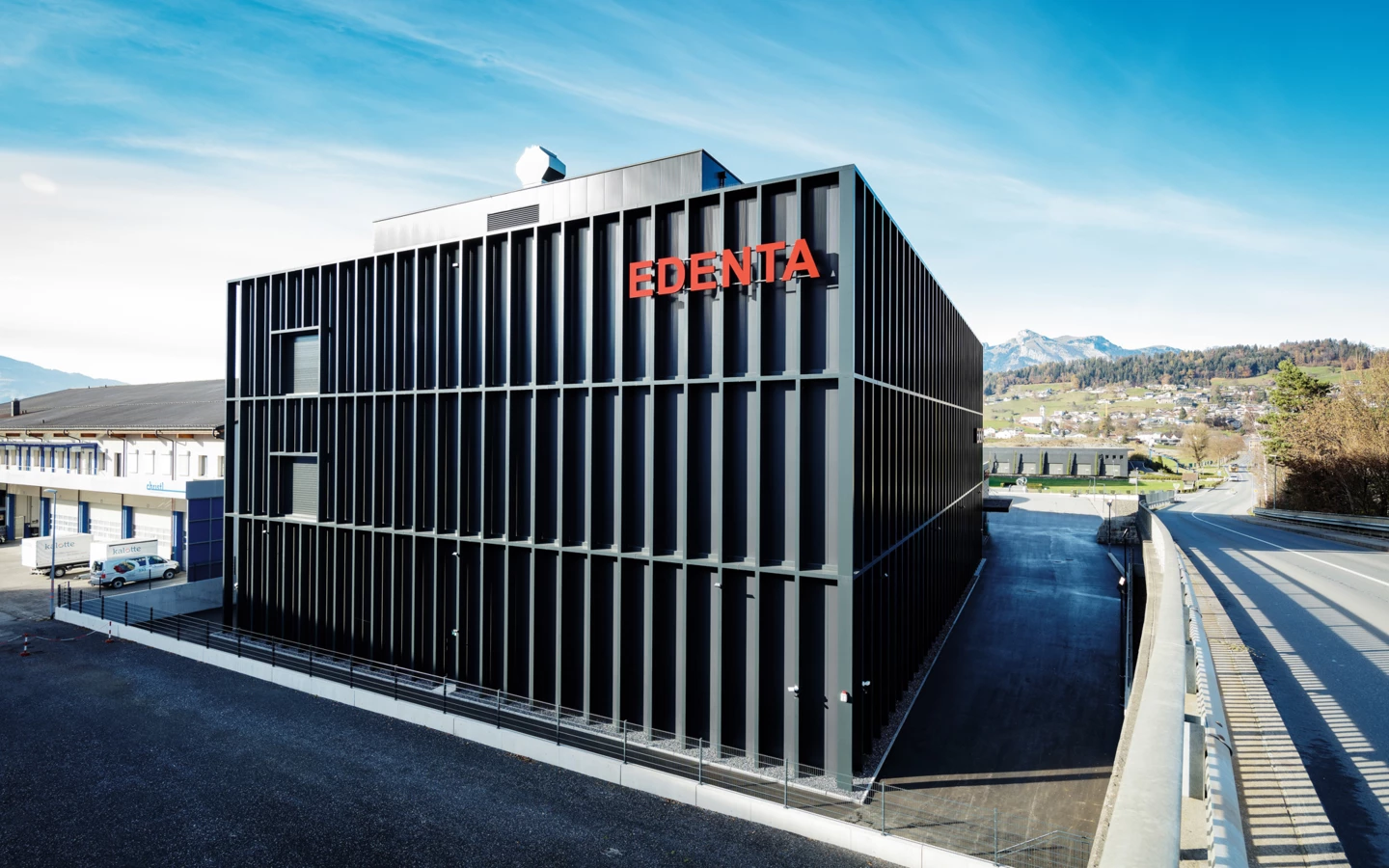 Modernes schwarzes Gebäude mit "EDENTA"-Schriftzug in Rot gegen einen blauen Himmel, daneben eine Straße und Berge.