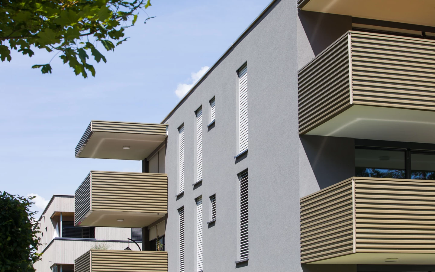 Graue Fassade der Wohnanlage Bahnhofstrasse mit mehreren Balkonen und eleganten Holzlatten-Brüstungen bei Tageslicht, umgeben von grünen Bäumen in Vandans (Bahnhofsstrasse)