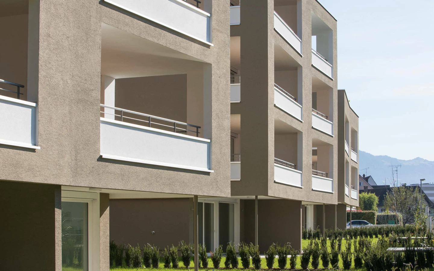 Moderne Wohnanlage Widum Lustenau mit beigefarbener Fassade, weißen Balkonen und grüner Gartenanlage unter blauem Himmel.