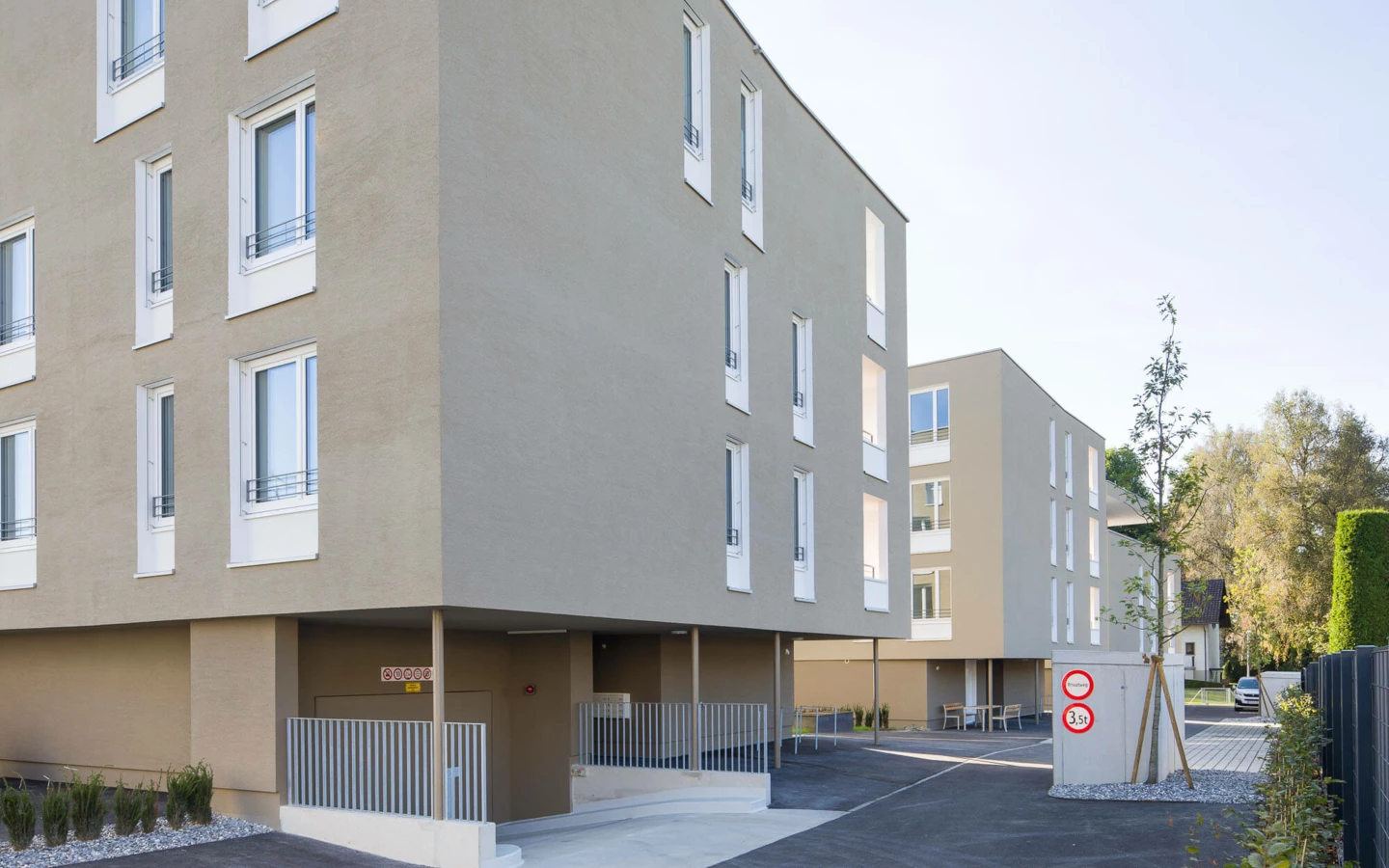 Moderne Wohnanlage Widum Lustenau 13 mit beige Fassaden, weiß umrahmten Fenstern, Tiefgarageneinfahrt und Straßenschildern.