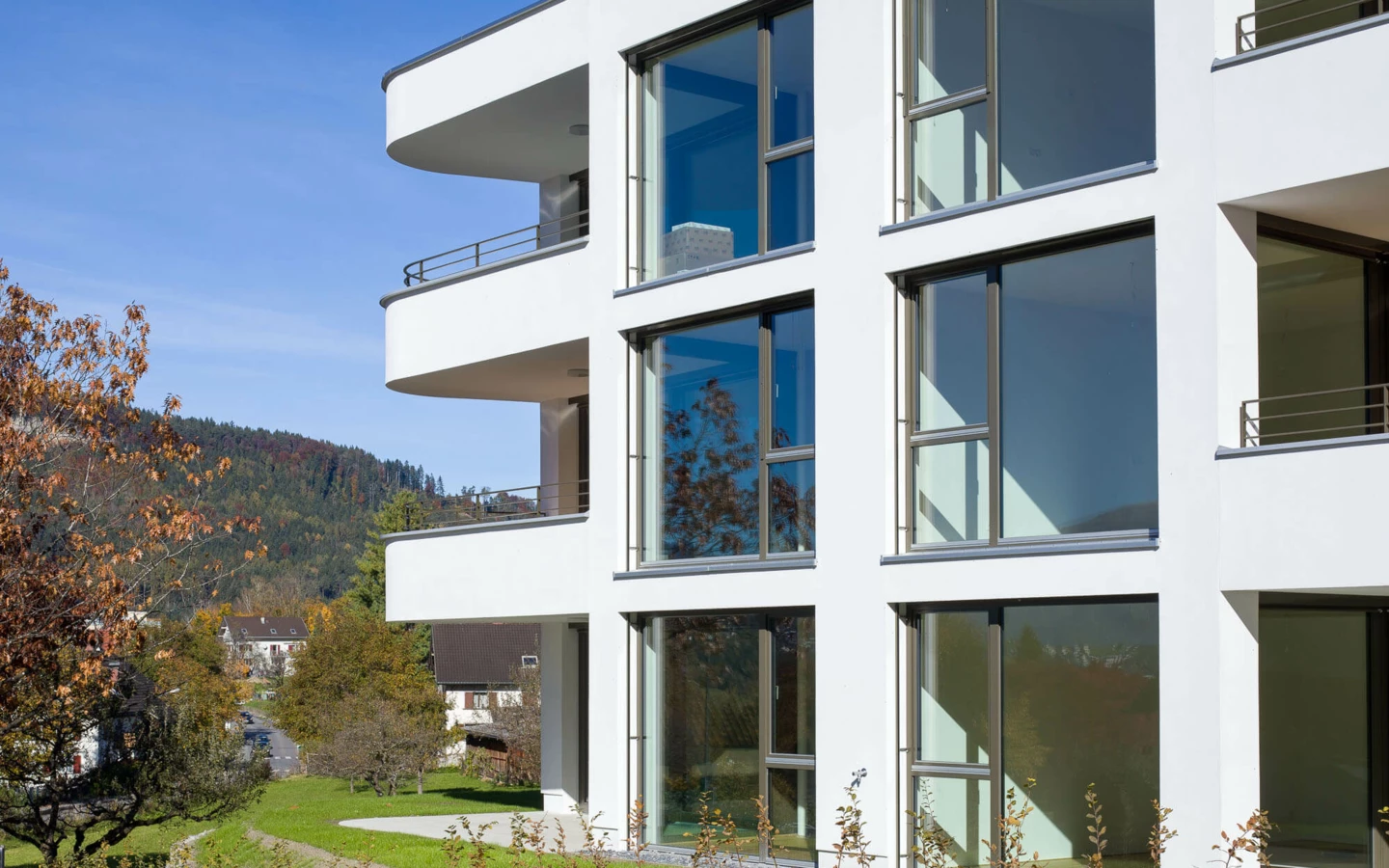 Moderne, weiße Wohnanlage in Feldkirch mit großen Fenstern und Balkonen vor einem bewaldeten Hügel an einem sonnigen Tag (Wolf-Huber-Straße).