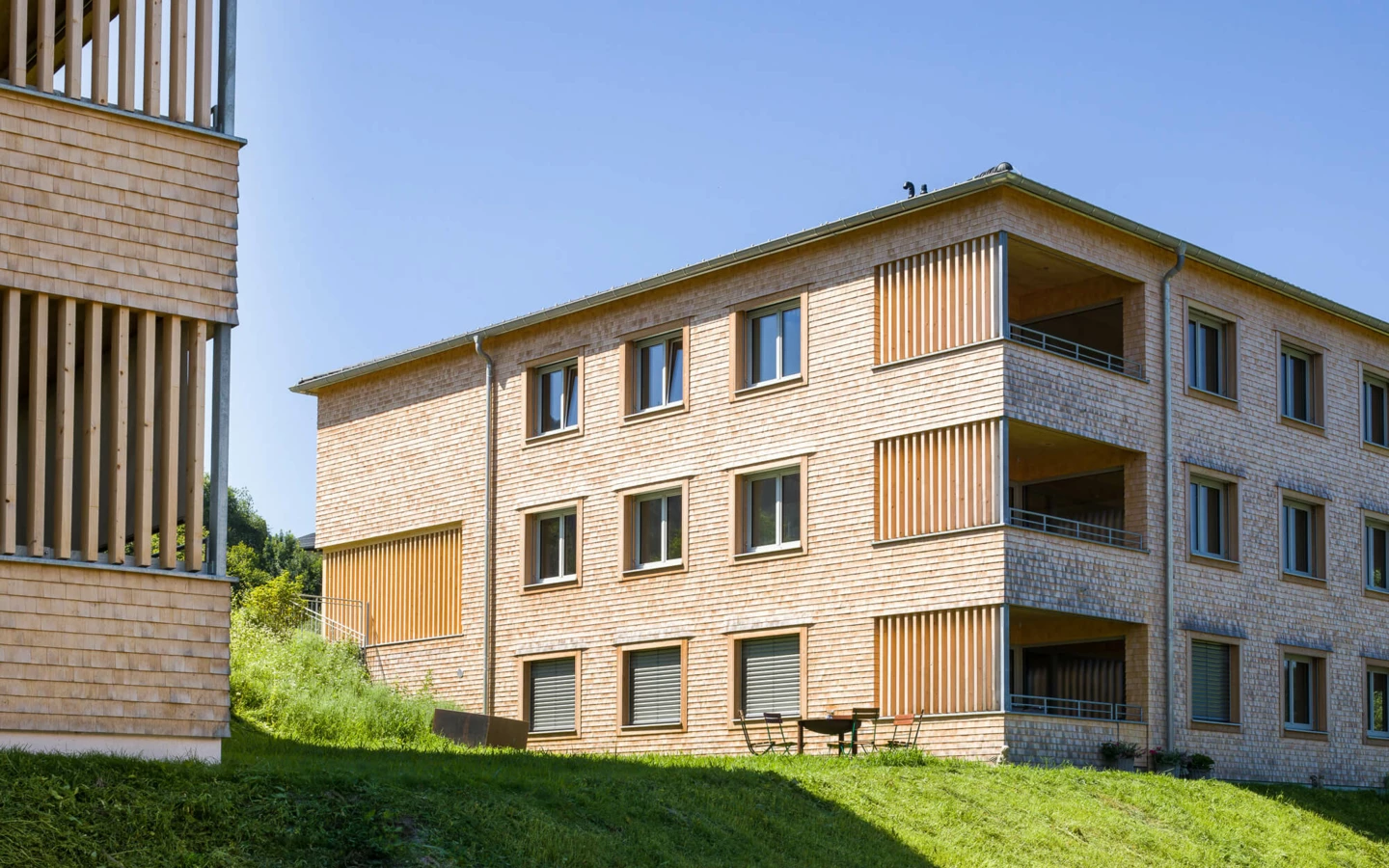 Zwei moderne Mehrfamilienhäuser mit Holzfassaden und Balkonen auf einem grünen Hügel in Alberschwende (Wohnanlage).