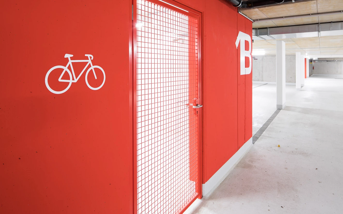 Roter Wandbereich in einer Tiefgarage mit Fahrradsymbol und "B" Markierung, daneben eine rote Gittertür zur Fahrradgarage (Baeumlequartier).