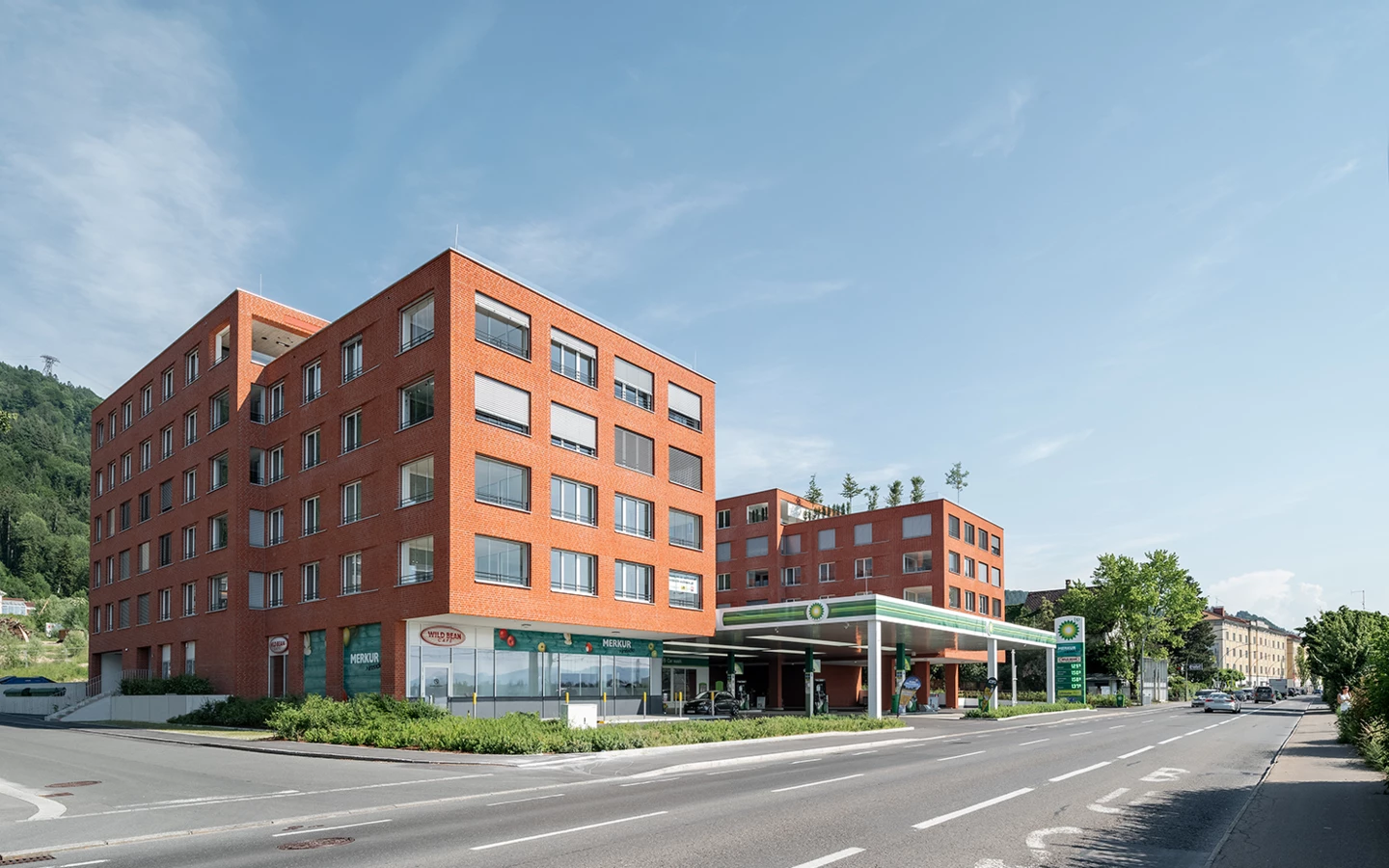 Zwei moderne Backsteingebäude neben einer BP-Tankstelle, mit "Merkur"- und "Wild Bean Cafe"-Beschriftungen, an einer Straße in Lochau (Seeblick).