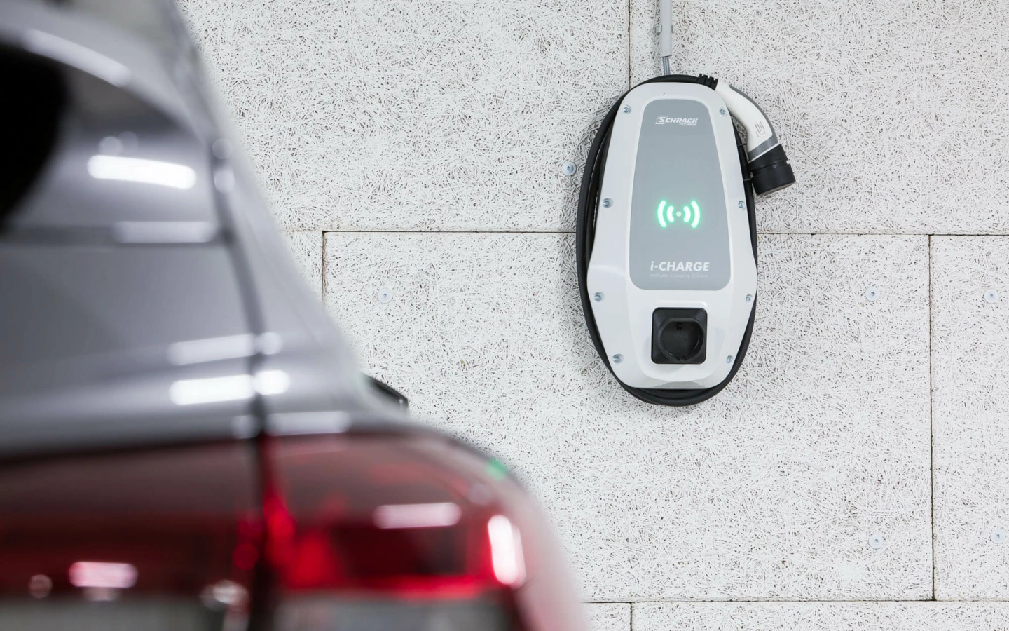Eine "i-CHARGE" E-Ladestation hängt an einer weißen Wand, ein dunkles Auto steht daneben (Wuhrbaumweg, Wohnanlage).