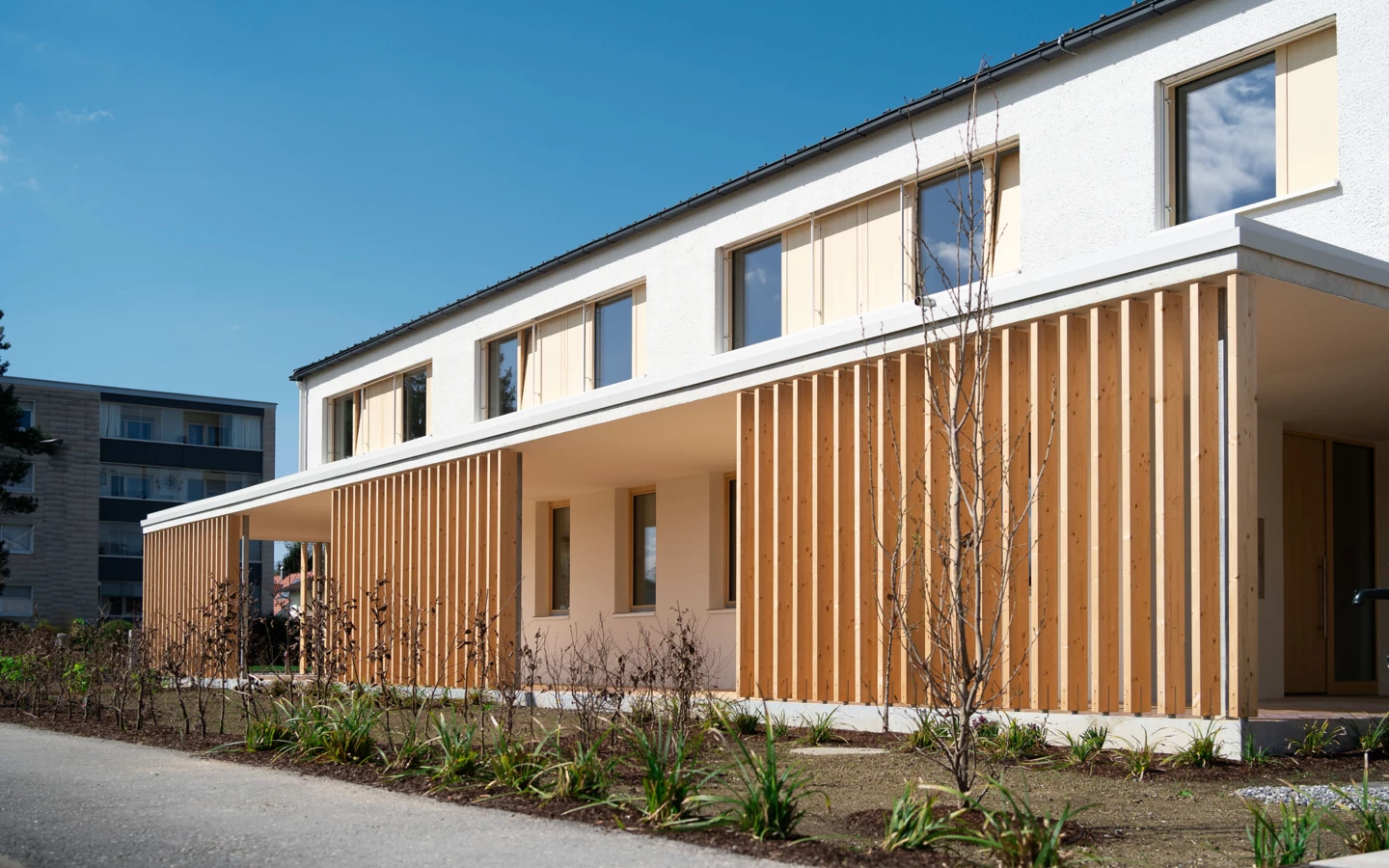 Modernes Wohngebäude mit weißer Fassade und vertikalen Holzlamellen als Sonnenschutz, daneben ein Fußweg und junge Bepflanzung (Wolfurt, Dieter).