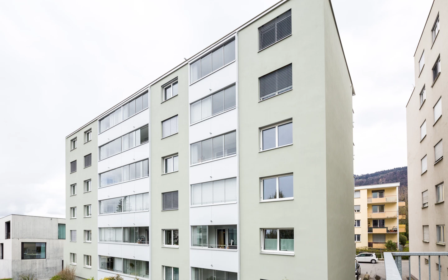 Modernes, mehrstöckiges Wohnhaus mit heller, grüngrauer Fassade und zahlreichen Fenstern, umgeben von weiterer Bebauung und Hügeln (Römerstraße).
