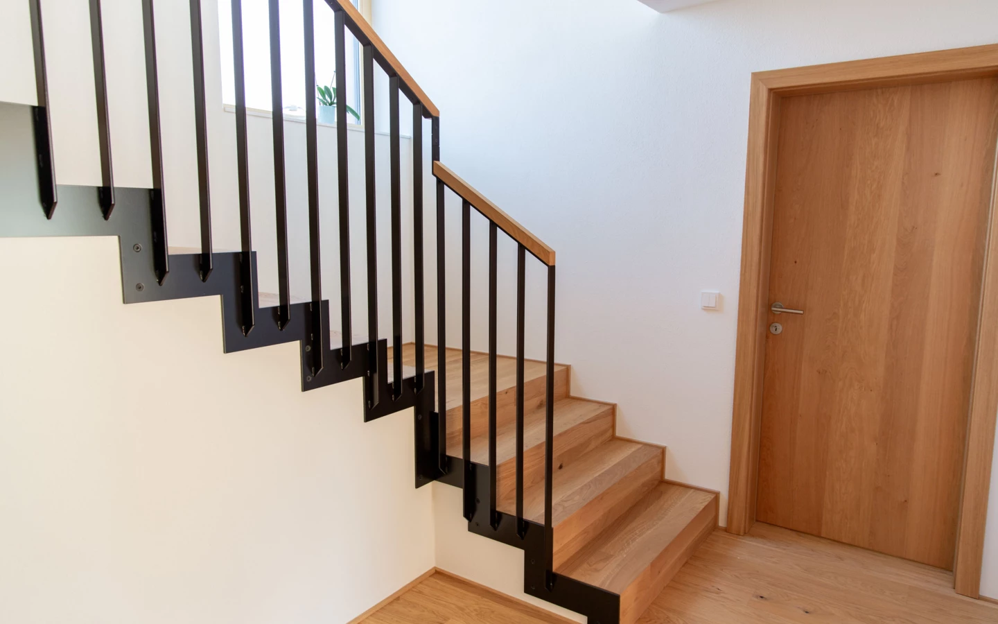 Holztreppe mit schwarzen Metallgeländern und heller Holztür in einem modern renovierten Einfamilienhaus in Dornbirn (Generalsanierung, Holzoptik).