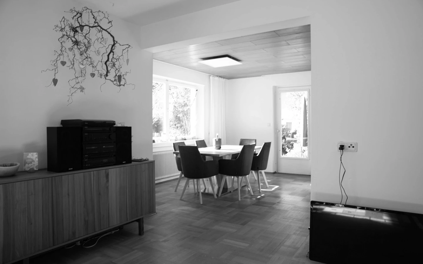 Wohnzimmer eines Einfamilienhauses mit Holzboden, Esstisch und Stühlen sowie einem Sideboard mit Musikanlage in Schwarz-Weiß (Generalsanierung, Holzoptik, Dornbirn).