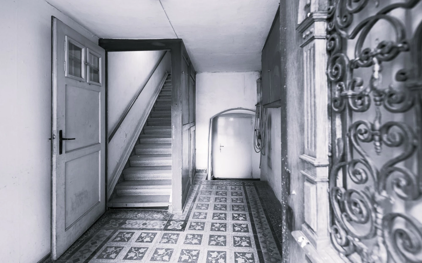 Schwarz-Weiß-Aufnahme eines alten Hausflurs mit schmiedeeisernem Geländer, gefliestem Boden und einer Treppe (Rathausstr).