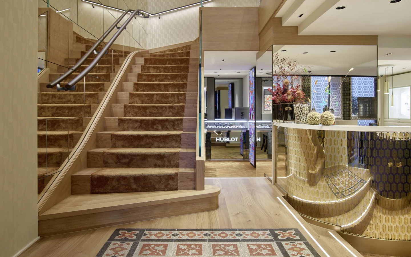Geschäftsinterieur mit Holztreppe mit braunem Teppich, gemusterter Fliesenboden und beleuchteter Vitrine mit Hublot-Schriftzug (Huber, Rathausstr).