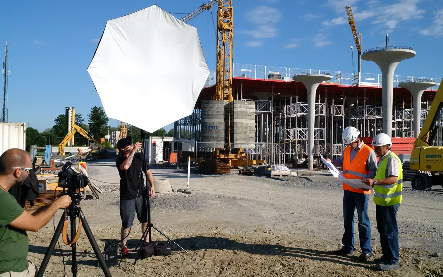 Fotoshooting auf einer Baustelle mit Kameramann, einem Lichttechniker und zwei Bauarbeitern, die Pläne begutachten.