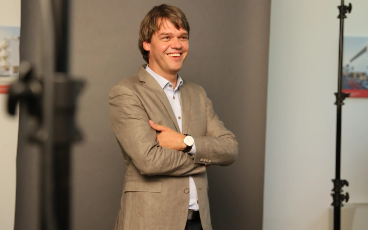 Ein lächelnder Mann in einem beigen Sakko und dunkler Hose steht mit verschränkten Armen vor einem grauen Hintergrund in einem Fotostudio.