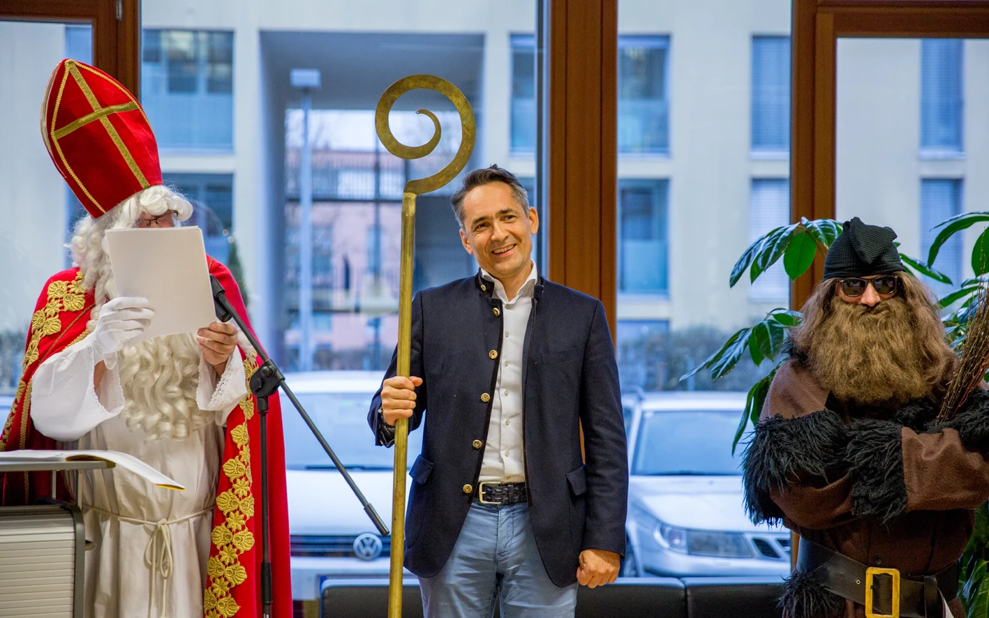 Ein Mann im Trachtenjanker hält einen Bischofsstab, flankiert von "Nikolaus" und "Knecht Ruprecht" vor einem Gebäude in Dornbirn (Hubert, Geburtstag).