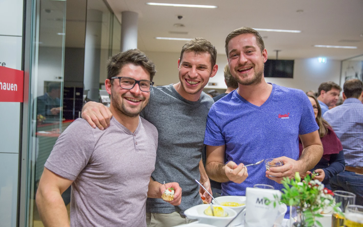 Drei lachende Männer stehen bei einer Veranstaltung vor einem Buffet mit kleinen Speisen und Getränken (Hubert, Geburtstag).