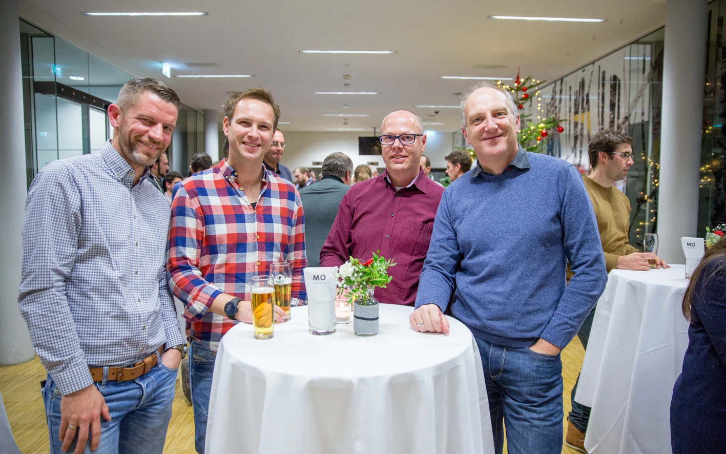 Vier lächelnde Männer unterhalten sich an einem Stehtisch mit Getränken bei einer Veranstaltung (Hubert, Geburtstag).