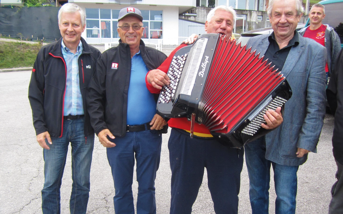 Four men standing outdoors, one playing a red and black Dallapè accordion, with buildings in the background (pensioners).