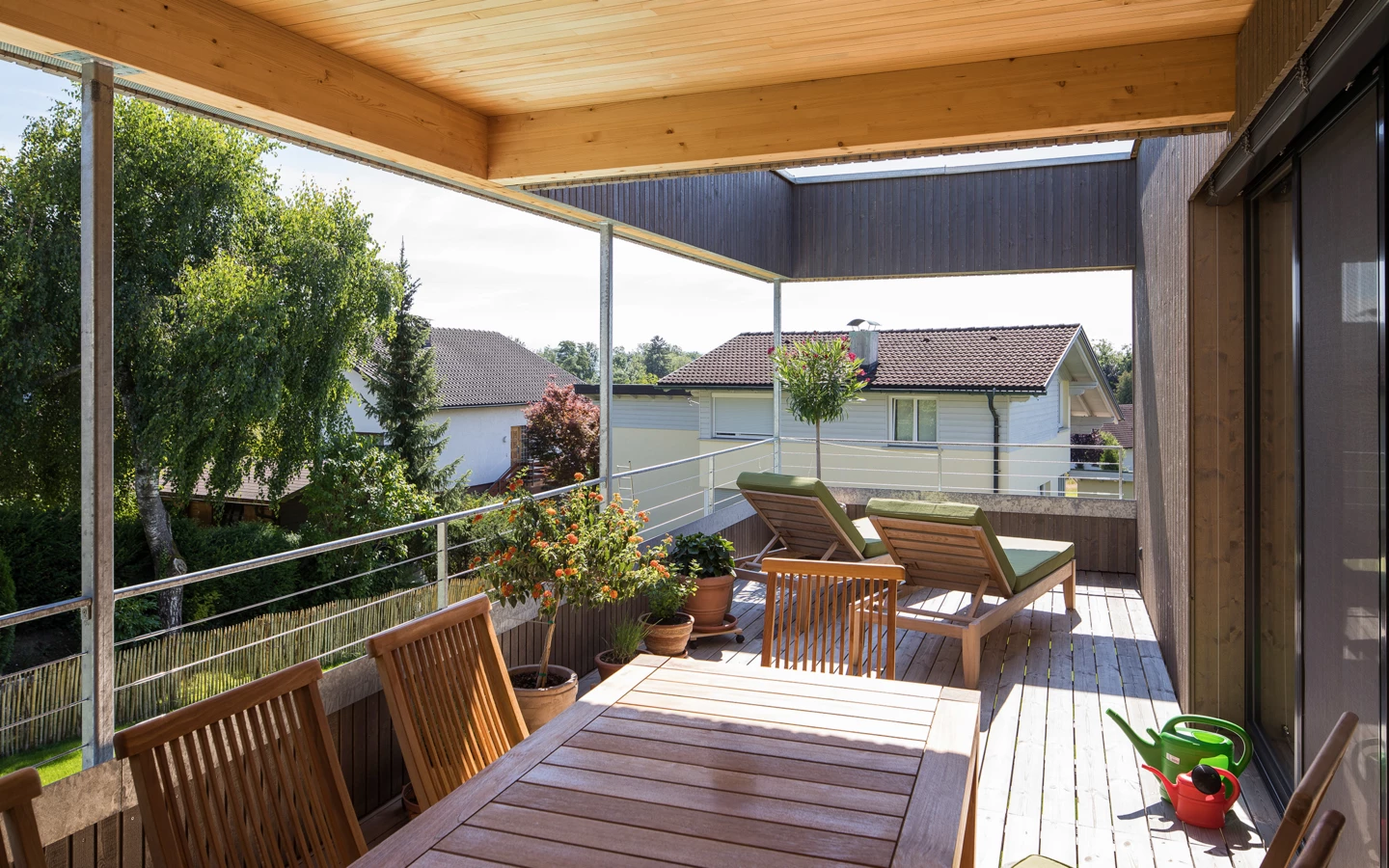Eine große Holzterrasse mit Esstisch, Stühlen und Sonnenliegen unter einem geschützten Dach, umgeben von Bäumen und Häusern (Hohenweiler).