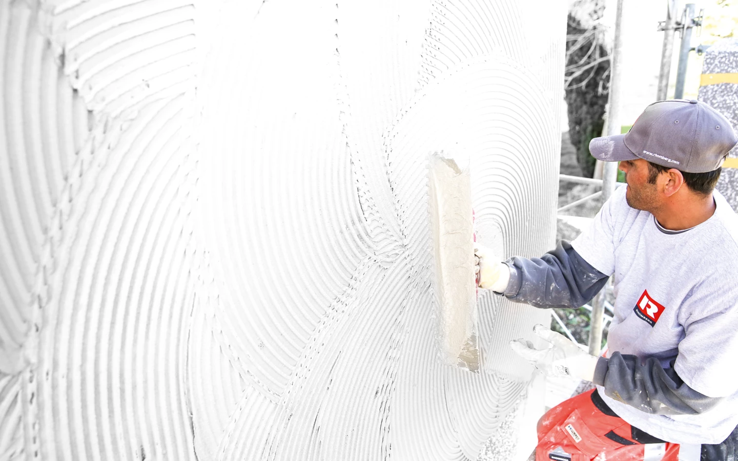 Ein Bauarbeiter in Arbeitskleidung trägt mit einer Glättkelle eine weiße Putzschicht auf eine Wand auf (Verputzarbeiten, Hochbau).