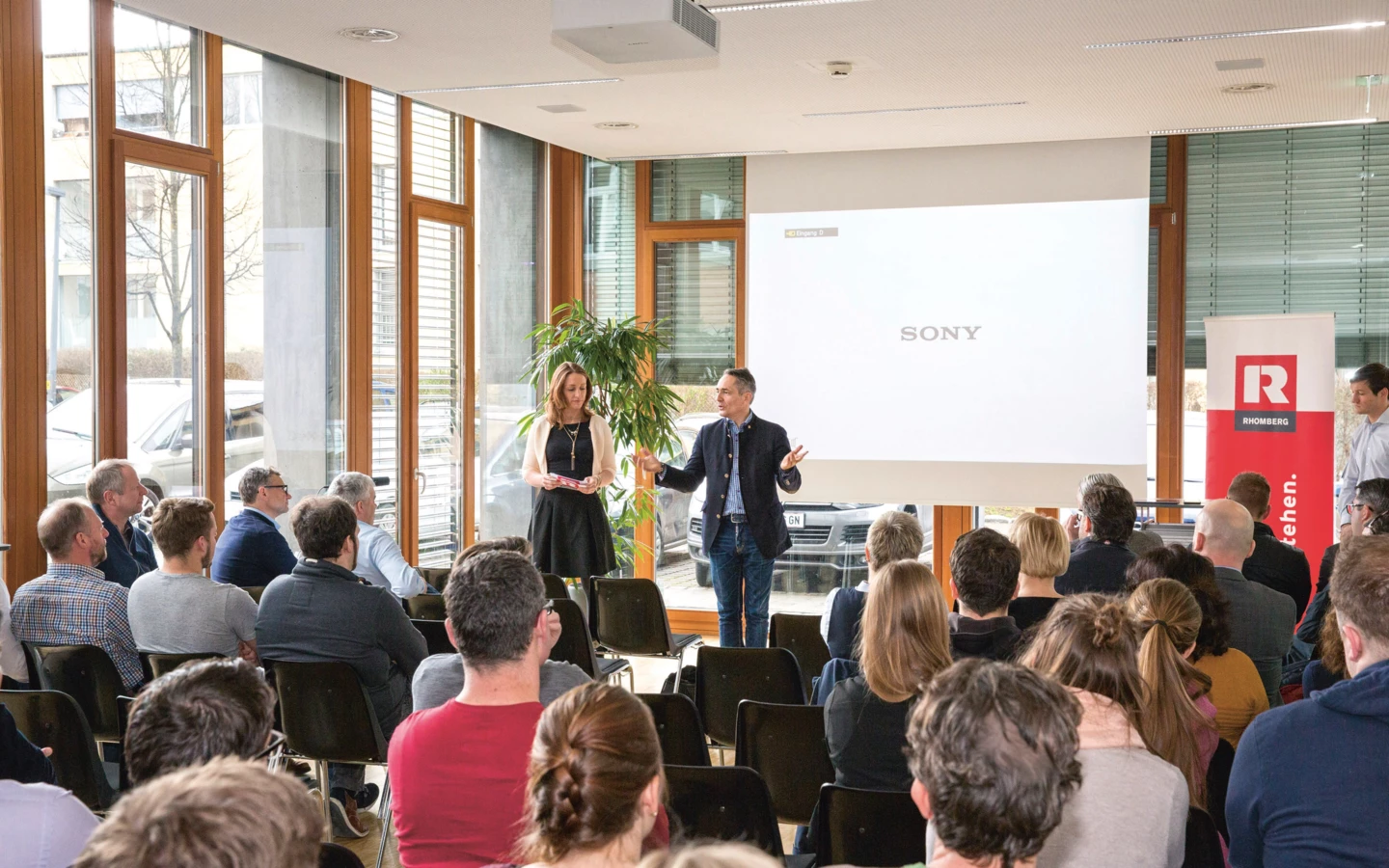 Zwei Personen präsentieren vor einem Publikum, mit Rhomberg Bau Flagge und Sony Logo an der Wand (Digital, Day).