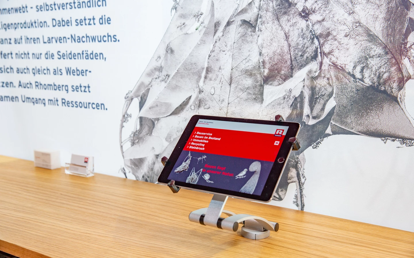Tablet mit Informationen zu Rhomberg Bau Leistungen auf Holztheke vor einer Wand mit Blattmotiven und Text.