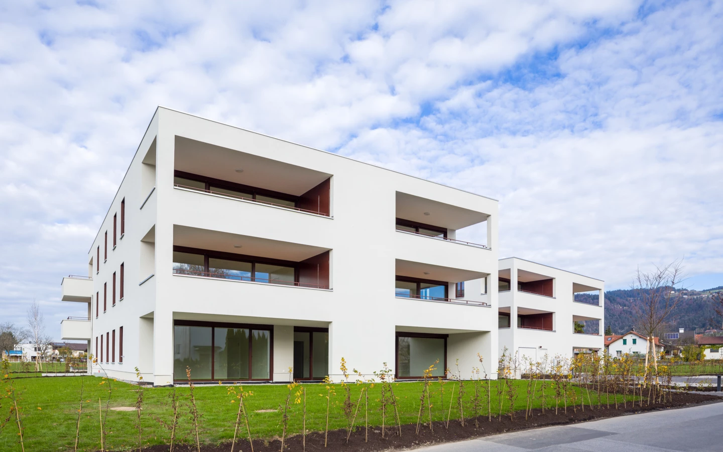 Zwei moderne weiße Wohngebäude mit Balkonen und großen Fenstern, umgeben von jungem Grün und Rasen, unter einem bewölkten Himmel (Unterhochstegstrasse).