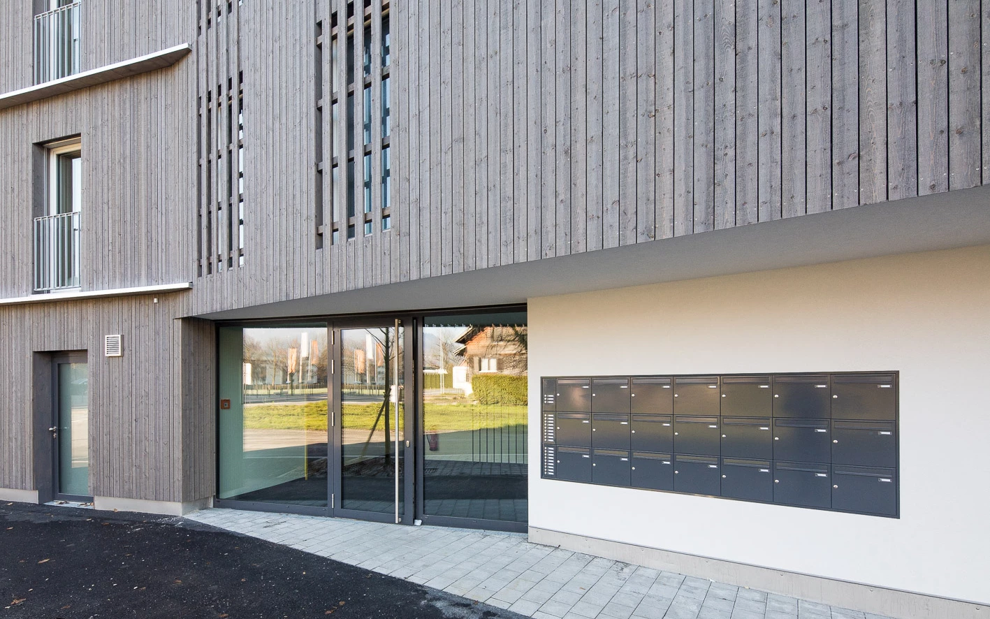 Eingangsbereich eines modernen Gebäudes mit Holzfassade, großen Glasfenstern und einer Reihe von Briefkästen an der weißen Wand (Froschweg).